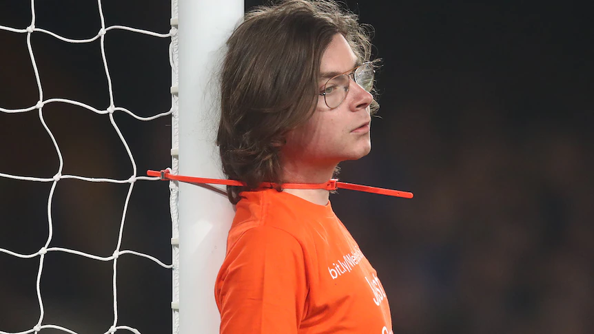 A man with long hair and glasses has a zip tie around his neck attached to a goalpost